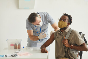Portrait of male doctor vaccinating young African American man wearing mask in medical clinic, copy...