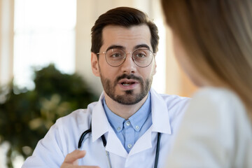 Obraz premium Head shot close up focused young male doctor in eyewear and medical uniform explaining healthcare treatment protocol to female patient, giving advices or prescribing medicine at checkup meeting.