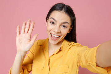 Close up young brunette attractive latin cute friendly woman 20s in yellow shirt do selfie shot on mobile phone waving hand say hi hello greeting isolated on pastel pink background studio portrait.