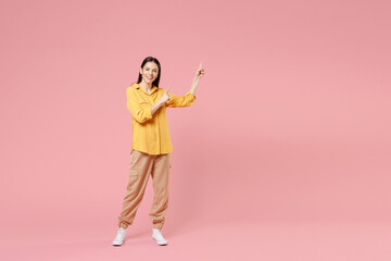Full length of young smiling brunette positive cute attractive latin woman 20s in yellow casual shirt point index finger aside on workspace area isolated on pastel pink background studio portrait.
