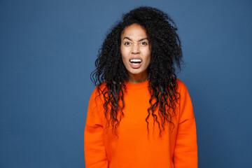 Shocked worried perplexed young african american woman wearing casual basic bright orange sweatshirt standing keeping mouth open looking camera isolated on blue color wall background studio portrait.