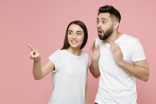 Young shocked surprised couple two friends bearded man woman in white basic blank print design t-shirt pointing finger aside on something say wow isolated on pastel pink background studio portrait