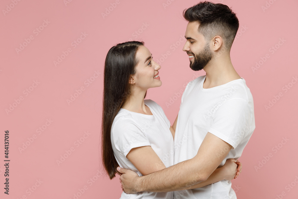 Wall mural Young happy couple in love two friends bearded man brunette woman 20s in white basic t-shirts looking to each other smiling hugging embracing isolated on pastel pink color background studio portrait.