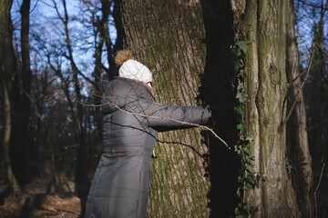 Waist-up portrait of warmly dressed woman hugging huge tree trunk in the woods.