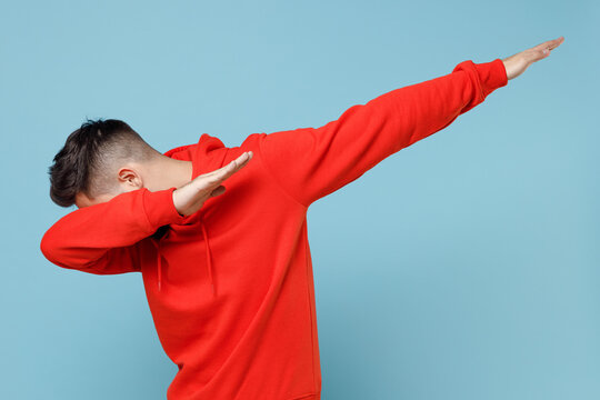 Young caucasian man 20s in casual red orange hoodie doing dab hip hop dance hands move gesture youth sign hiding covering face isolated on blue background studio portrait People lifestyle concept.