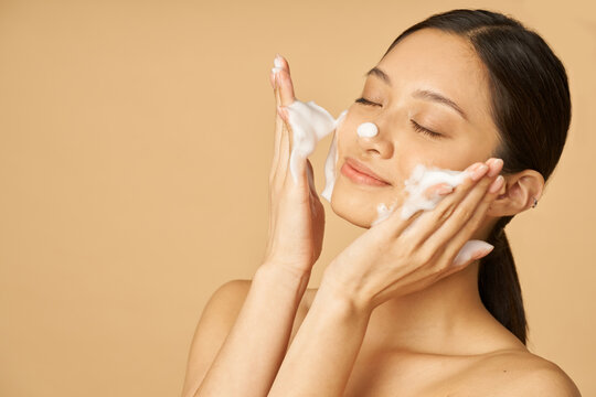 Beauty Portrait Of Happy Young Woman Smiling With Eyes Closed While Applying Gentle Foam Facial Cleanser Isolated Over Beige Background