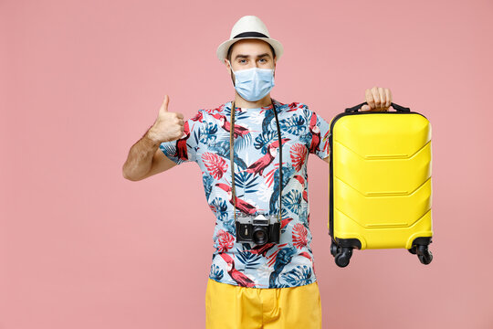 Cheerful young traveler tourist man in sterile mask to safe from coronavirus hold suitcase showing thumb up isolated on pink background. Passenger traveling on weekend. Air flight journey concept.