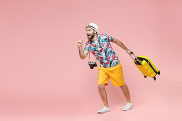 Full length of shocked young traveler tourist man in summer clothes hat running hold suitcase isolated on pink background studio portrait. Passenger traveling on weekends. Air flight journey concept.