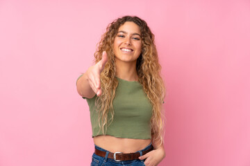 Young blonde woman with curly hair isolated on pink background shaking hands for closing a good deal