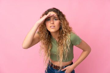 Young blonde woman with curly hair isolated on pink background looking far away with hand to look something