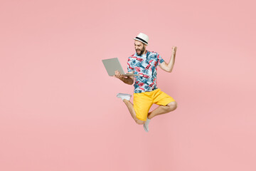 Full length happy traveler tourist man in summer clothes hat jump work on laptop computer doing winner gesture isolated on pink background. Passenger traveling on weekend. Air flight journey concept.