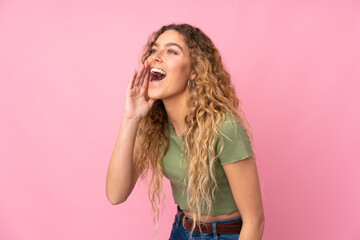 Young blonde woman with curly hair isolated on pink background shouting with mouth wide open to the lateral