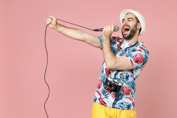 Laughing young traveler tourist man in summer clothes hat photo camera sing song in microphone isolated on pink background studio portrait. Passenger traveling on weekend. Air flight journey concept.