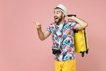 Amazed young traveler tourist man in summer clothes hat photo camera hold suitcase pointing index...