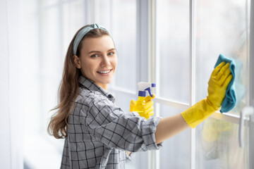 Smiling young woman with detergent and rag cleaning window glass. Professional sanitary service...