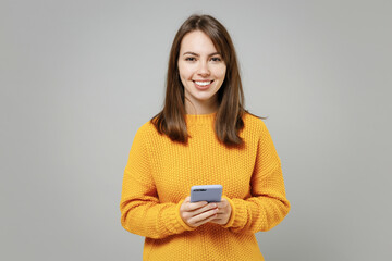 Young smiling cheerful woman 20s wearing casual knitted yellow sweater hold in hand using mobile cell phone chatting browsing typing sms messaging isolated on grey color background studio portrait. - Powered by Adobe