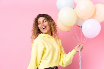 Young blonde woman with curly hair catching many balloons isolated on pink background