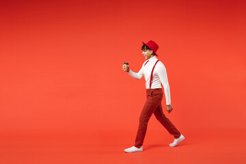 Fototapeta na wymiar Full length side view of young spanish latinos smiling fashionable man 20s in hat white shirt trousers, suspenders hold paper cup of coffee drink walking isolated on red background studio portrait.