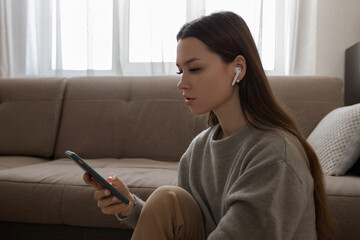 A young girl sits in plain clothes on the floor next to a brown sofa. The girl in wireless headphones looks at the phone. The girl is talking on video communication in headphones. The girl listens to