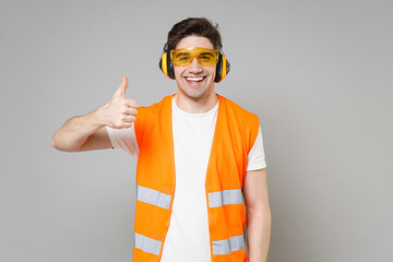 Young employee happy handyman man in orange vest protective ear muffs look camera show thumb up gesture isolated on grey background studio. Instruments for renovation apartment. Repair home concept.
