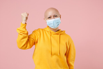 Young bald excited woman 20s without hair in yellow casual shirt in sterile face mask to safe coronavirus virus covid-19, pandemic quarantine do winner gesture clench fist isolated on pink background.