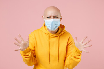 Young bald angry indignant woman 20s without hair wear yellow casual shirt in sterile face mask to safe from coronavirus virus covid-19, pandemic quarantine spread hands isolated on pink background