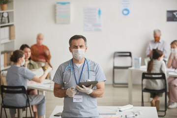Fototapeta premium Waist up portrait of mature male doctor wearing mask and looking at camera while working at vaccination center, copy space