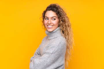 Young blonde woman with curly hair wearing a turtleneck sweater isolated on yellow background with arms crossed and happy