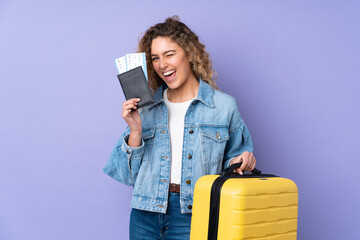 Young blonde woman with curly hair isolated on purple background in vacation with suitcase and passport