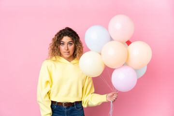Young blonde woman with curly hair catching many balloons isolated on pink background having doubts and with confuse face expression