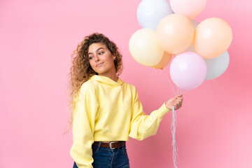 Young blonde woman with curly hair catching many balloons isolated on pink background