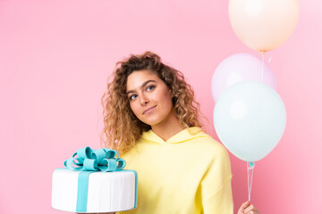 Young blonde woman with curly hair catching many balloons and holding a big cake isolated on pink background
