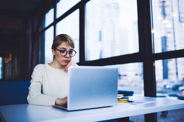 Pensive woman in eyeglasses using laptop