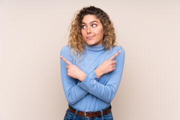 Obraz premium Young blonde woman with curly hair wearing a turtleneck sweater isolated on beige background pointing to the laterals having doubts