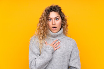 Young blonde woman with curly hair wearing a turtleneck sweater isolated on yellow background surprised and shocked while looking right