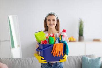 Joyful young maid holding bucket full of cleaning supplies at living room