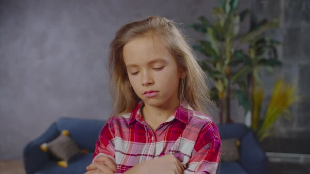 Portrait of cute upset pouting elementary age girl with long blonde straight hair standing with arms crossed, looking with sad expression, feeling disappointment, stress and helpless in home interior