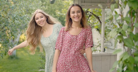 Two young beautiful girls wearing dresses posing on the porch, smiling, dancing, looking at camera, sunny backyard garden view, summer time in the countryside.