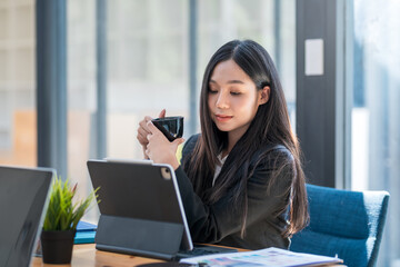 Asian businesswoman sitting at a desk in the office drinking coffee look at a tablet.