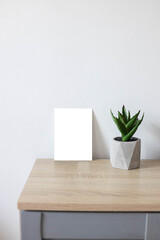 White empty paper blank frame mockup on home wooden table. Modern grey concrete flowerpot with succulent plant. White wall background