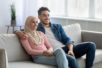 Obraz premium Cheerful muslim couple sitting on couch at home, copy space