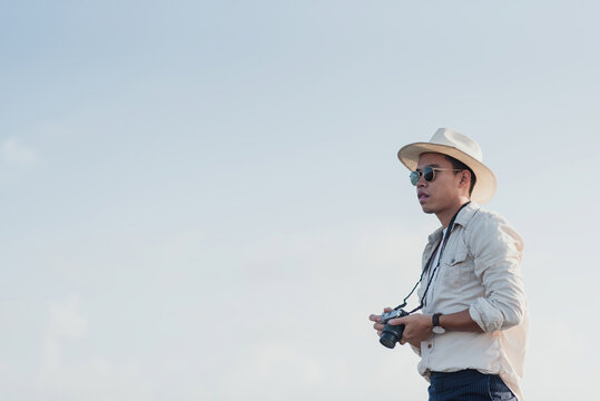 Traveler Concept: An Asian man holding a camera in his phone looking forward to take a picture against a white sky background.