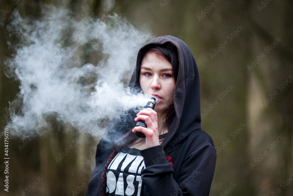 Wall mural Vape teenager. Young pretty caucasian brunette girl with problem skin in the hood smoking an electronic cigarette on the street in the spring. Deadly bad habit.