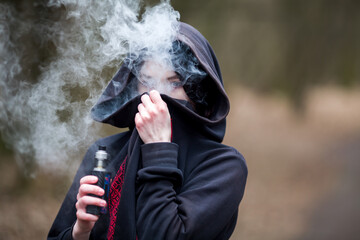 Vape teenager. Young pretty caucasian brunette girl with problem skin in the hood smoking an...