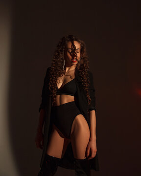 young woman with long curly hair  in black lingerie and jacket, in lacquered hessian boots is standing  near white wall background with shadows  in photo studio, fashion concept, free space