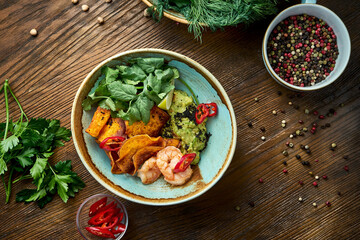 Appetizing mexican appetizer - avocado guacamole with shrimps, vegetable chips and cilantro in a bowl on a wooden background. Close up view