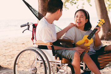 Obraz premium Asian special child on wheelchair is singing, playing ukulele happily on the beach with parent,Natural sea beach background,Life in the education age of disabled children,Happy disability kid concept.
