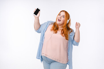 Corpulent ginger-haired young woman listening to the music on the phone isolated over white background
