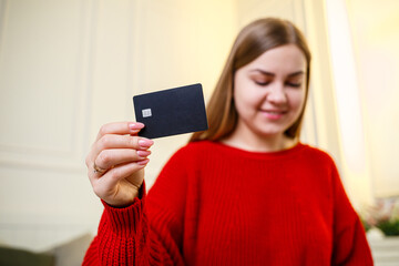 Naklejka premium Young beautiful woman in a red sweater holding a bank card. Credit card for online purchases. Selective focus