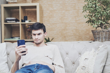 Indignant young man embarrassed looking at screen of his phone while sitting on couch at home